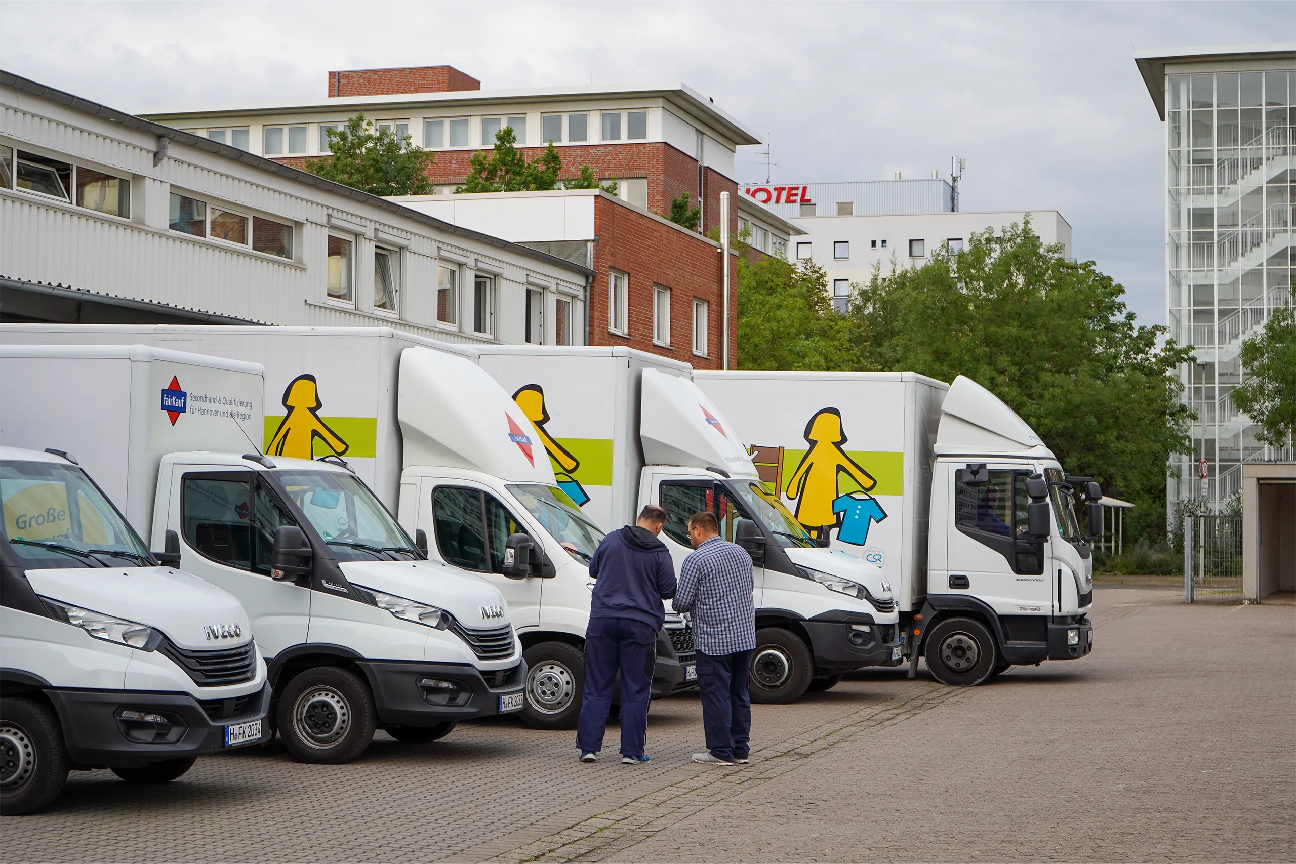Zwei Männer stehen zwischen mehreren weißen Lieferwagen auf einem Parkplatz vor einem Bürogebäude.