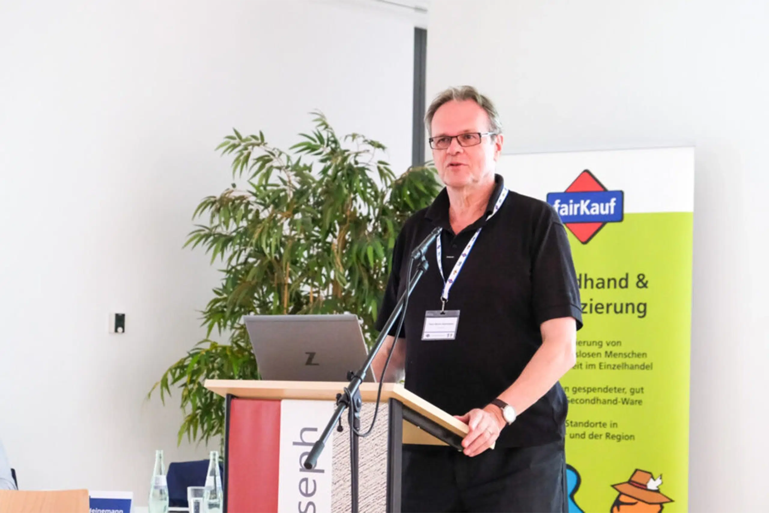 Person hält einen Vortrag am Redepult, mit Laptop und fairKauf-Banner im Hintergrund.