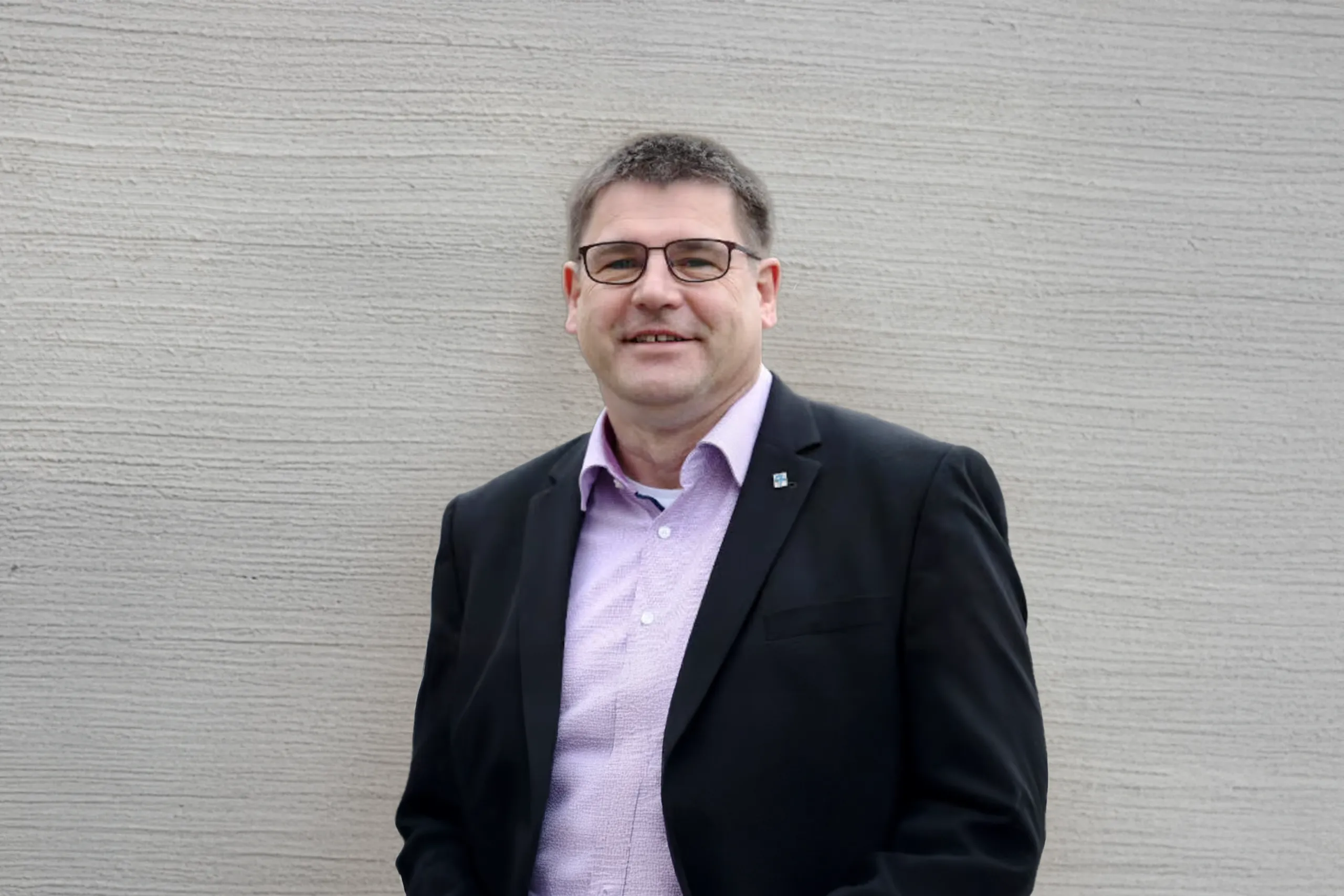 Person mit kurzer grauer Frisur, Brille und dunklem Sakko steht draußen und lächelt in die Kamera.