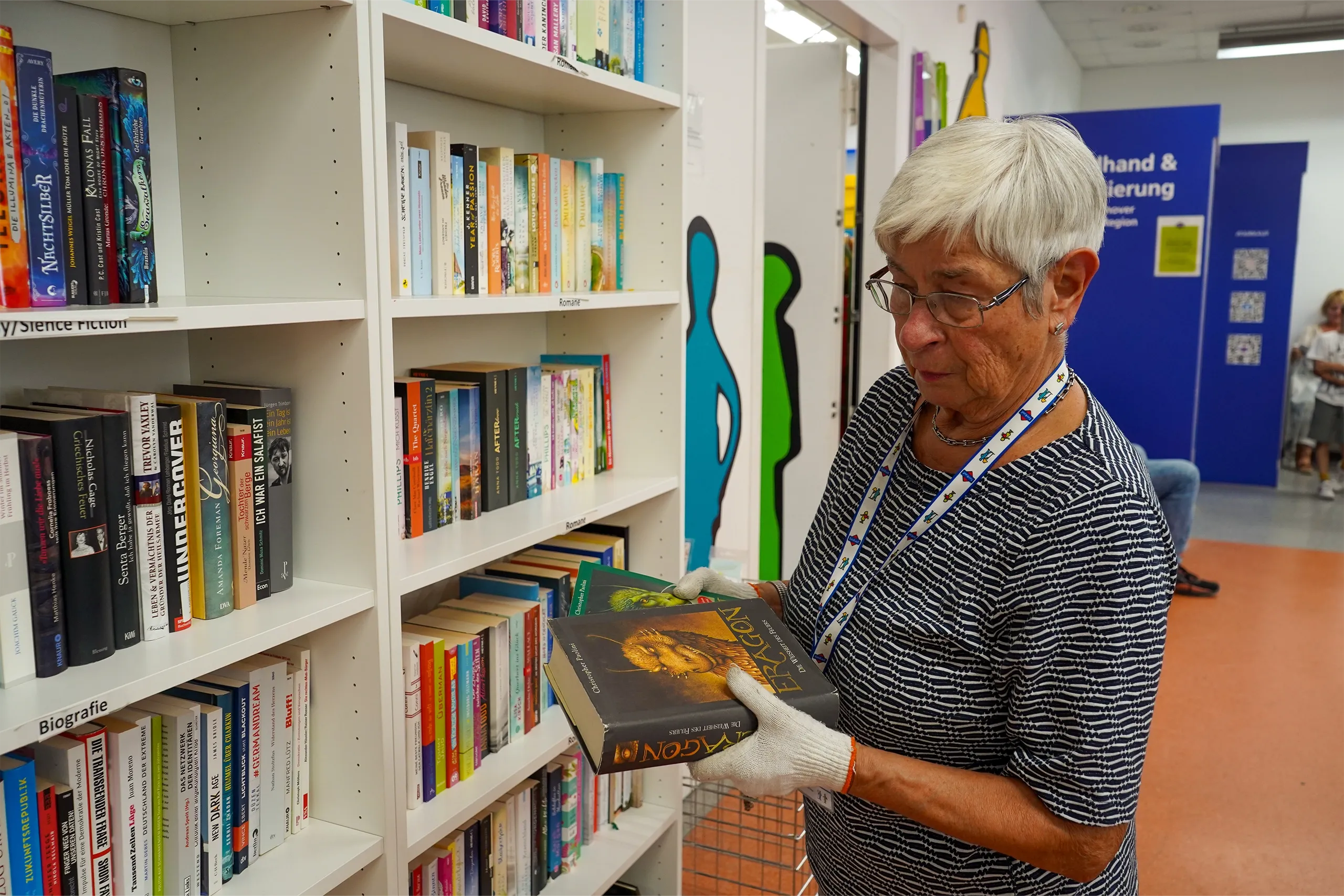 Eine ältere Frau mit grauen Haaren und Brille hält zwei Bücher in den Händen und schaut auf ein Bücherregal in einer Bücherei.
