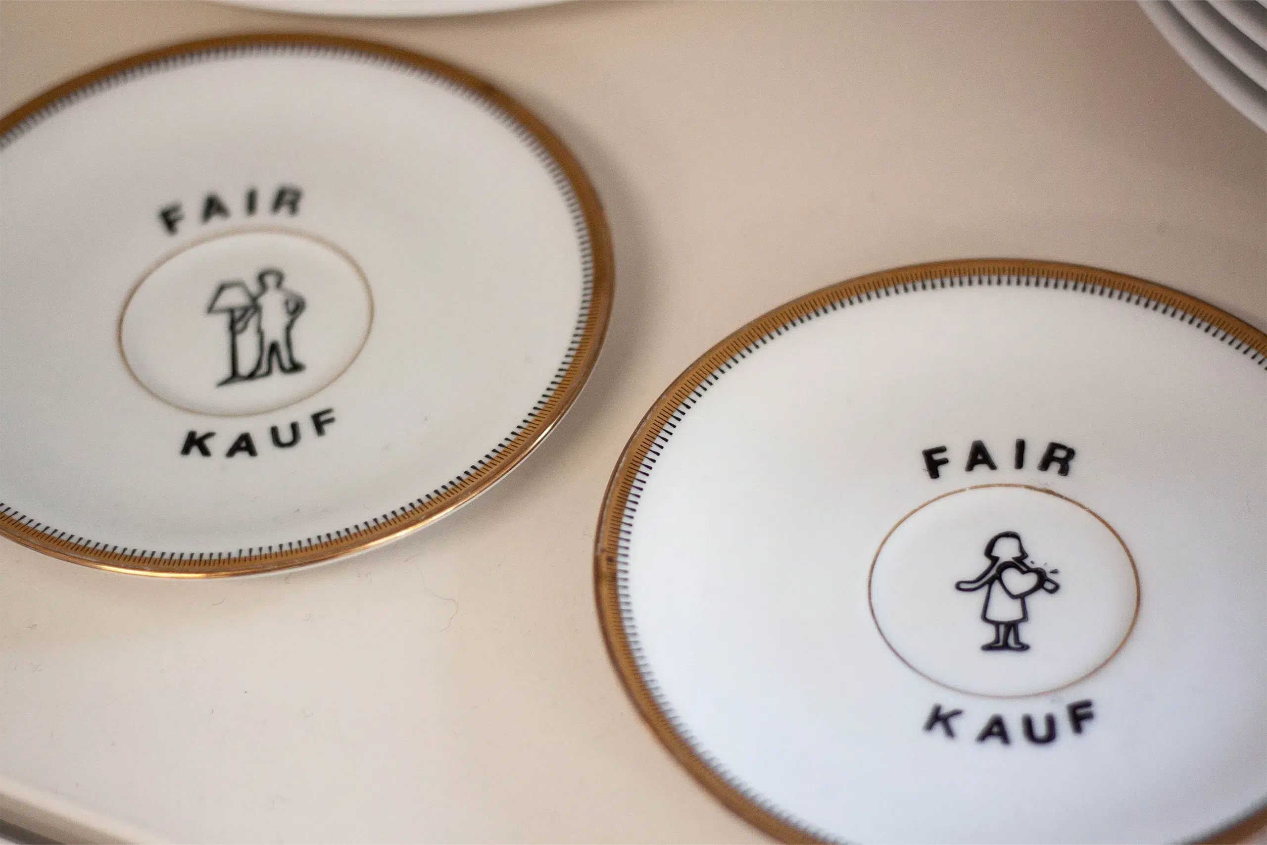 Zwei Untertassen mit dem Aufdruck "FAIR KAUF" und symbolischen Darstellungen von Personen.