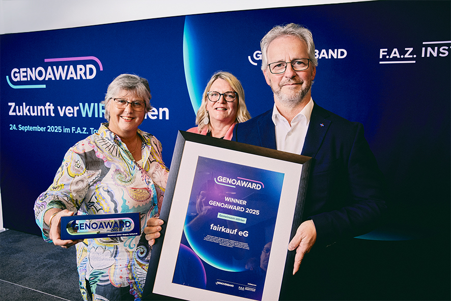 Drei Personen präsentieren den GENOAWARD 2025 für fairkauf eG vor einer Sponsorentafel.